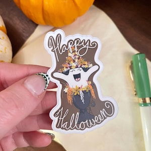 May include: A white sticker with a cartoon ghost design. The ghost has a smiling face and is wearing a flower arrangement on its head. The words "Happy Halloween" are written in a decorative font.
