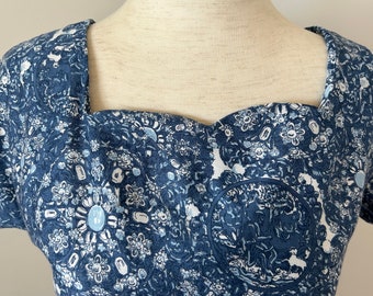 May include: A blue and white patterned top with a square neckline. The fabric features a detailed floral and medallion design. The top has short sleeves and a fitted bodice, showcasing a vintage aesthetic.