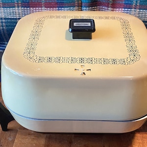 Op de afbeelding: Crèmekleurige elektrische braadpan met een zwarte handgreep en een decoratieve zwarte bloemenrand op het deksel. De braadpan staat op een houten dienblad met zwarte steunen. Een kruisvormige ventilatieopening is zichtbaar.