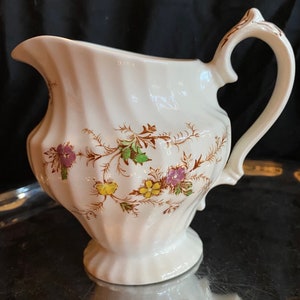 Puede incluir: Una jarra de crema de cerámica blanca con un diseño floral en tonos marrones, verdes, amarillos y morados. La jarra tiene un cuerpo acanalado y un asa.