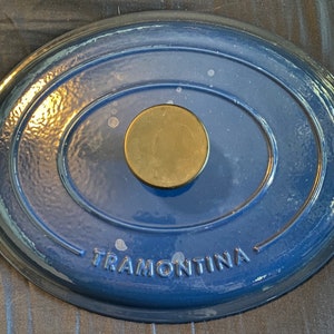 Puede incluir: Tapa de olla ovalada de hierro fundido esmaltado azul con pomo de latón y la palabra "Tramontina" inscrita en la parte inferior.