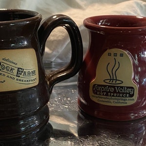 Puede incluir: Dos tazas de cerámica con asas. La taza marrón tiene una etiqueta que dice "Sand Rock Farm Bed and Breakfast Est. 1887 Aptos, CA." La taza burdeos tiene una etiqueta que dice "Surprise Valley Hot Springs surprisevalleyhotsprings.com Cedarville, California."