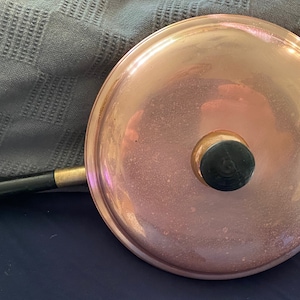 Puede incluir: Tapa de olla de cobre con un pomo negro y un mango de madera. La tapa es brillante y tiene una ligera pátina.