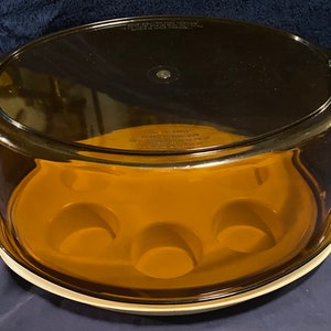 Puede incluir: Una cúpula para tartas de vidrio transparente con una base marrón y un borde blanco. La base tiene cuatro hendiduras para sostener cupcakes u otras golosinas.