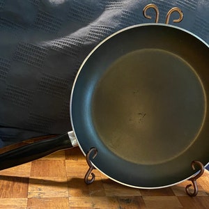 May include: A black non-stick frying pan with a black handle and a silver rim. The pan is sitting on a wooden stand with two metal hooks.