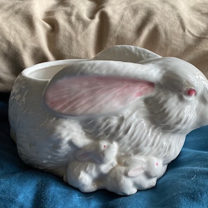 Könnte beinhalten: Weißer Keramik-Pflanztopf in Form eines Kaninchens mit rosa Ohren und drei kleineren Kaninchen, die sich an seinen Füßen kuscheln.