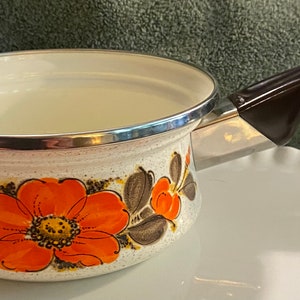 May include: A white enamel saucepan with a brown handle and a floral design featuring orange, brown, and white flowers.