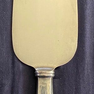 May include: A silver cake server with a hammered handle and an ornate design at the base. The server has a smooth, oval-shaped blade.