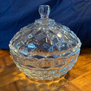 Puede incluir: Un cuenco de cristal transparente con tapa y un patrón de diamantes. El cuenco redondo tiene una tapa a juego con un pequeño asa decorativa. El cristal refleja la luz, creando un efecto brillante. El cuenco es probablemente vintage.