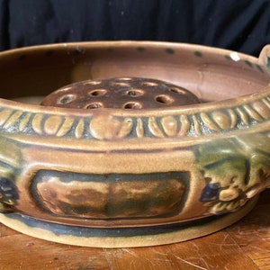 May include: A brown and green ceramic planter with a decorative floral design and a central hole for a plant. The planter has two handles on the sides.