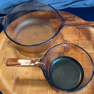 Puede incluir: Dos piezas de cocina de vidrio marrón con bordes azules. La pieza más grande tiene un asa y un fondo redondo. La pieza más pequeña tiene un asa y un interior antiadherente negro.