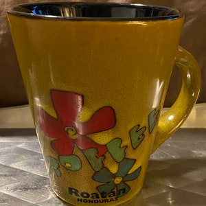 May include: A yellow ceramic coffee mug with a colorful floral design and the text "Roatan Honduras".