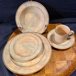 May include: A set of four ceramic dinnerware pieces in a light beige color with a marbled pattern. The set includes a large bowl, a smaller bowl, a dinner plate, and a teacup and saucer. The pieces have a thin black trim around the edges.