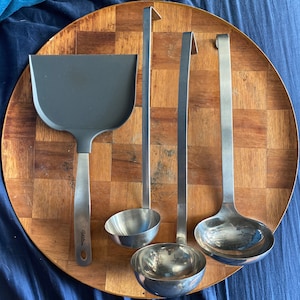 May include: A set of four kitchen utensils on a wooden surface. The set includes a spatula, a ladle, and two ladles with long handles. The spatula has a black handle with the word "Williams" printed on it. The ladles are made of stainless steel.