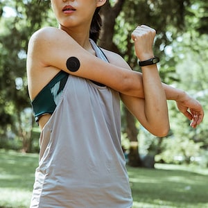 May include: A woman wearing a gray tank top and a black fitness tracker on her wrist stretches her arm. A black round sensor is attached to her upper arm.