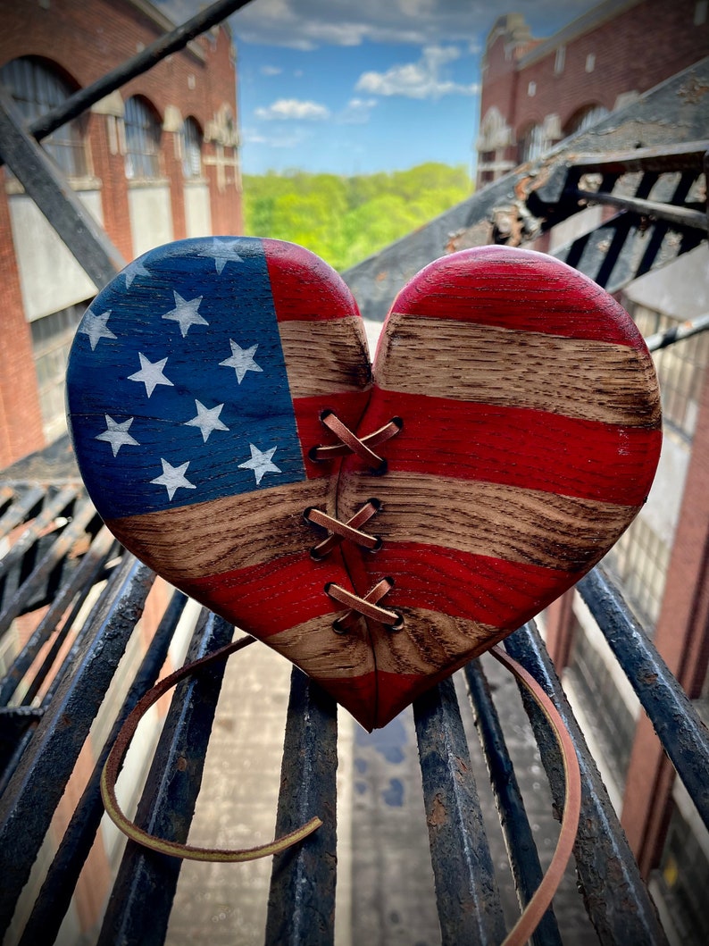 May include: A wooden heart-shaped ornament painted with the American flag design. The heart is stitched together with brown leather cord.