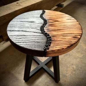 Op de afbeelding: Een ronde houten kruk met een zwart en naturel houten blad. Het blad is in twee&euml;n gedeeld met een zwarte leren strook in het midden. De kruk heeft een zwarte metalen basis.
