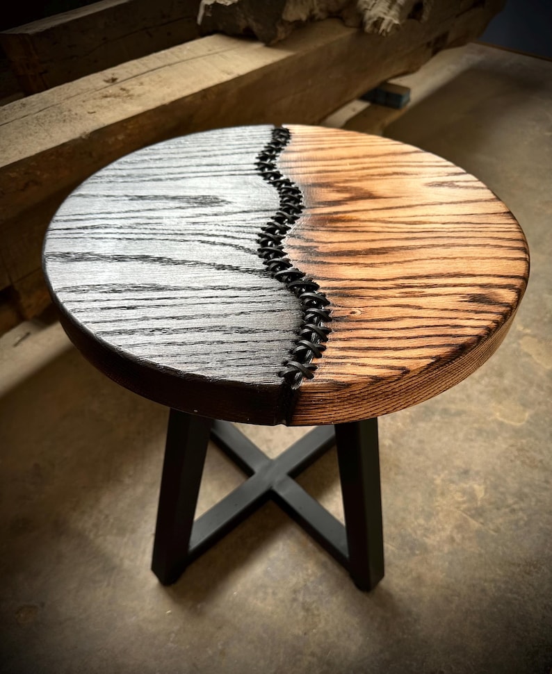 Op de afbeelding: Een ronde houten kruk met een zwart en naturel houten blad. Het blad is in twee&euml;n gedeeld met een zwarte leren strook in het midden. De kruk heeft een zwarte metalen basis.