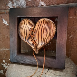 Puede incluir: Una escultura de corazón de madera con un acabado de grano de madera carbonizado oscuro, que presenta una división vertical unida por cordones de cuero fino de color tostado. El corazón está montado dentro de un marco de madera cuadrado teñido de oscuro, con dos largos cordones de cuero colgando.