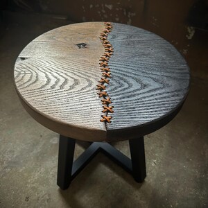 Op de afbeelding: Een ronde houten kruk met een zwarte metalen basis. De bovenkant van de kruk is in twee&euml;n gesplitst, met &eacute;&eacute;n kant donkerbruin en de andere kant lichtgrijs gebeitst. De twee helften worden bij elkaar gehouden met een leren stiksel.