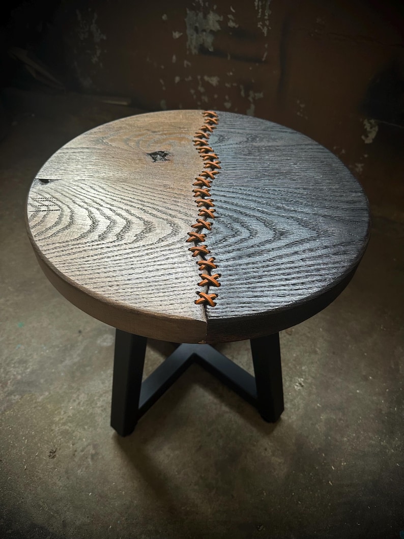 Op de afbeelding: Een ronde houten kruk met een zwarte metalen basis. De bovenkant van de kruk is in twee&euml;n gesplitst, met &eacute;&eacute;n kant donkerbruin en de andere kant lichtgrijs gebeitst. De twee helften worden bij elkaar gehouden met een leren stiksel.