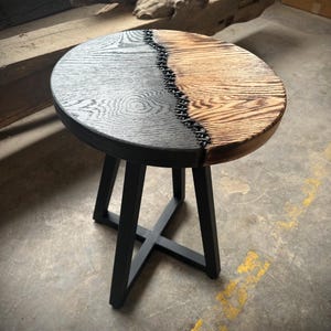 Op de afbeelding: Een ronde houten bijzettafel met een zwart en naturel houten blad. Het blad is in twee&euml;n gedeeld met een zwarte epoxy naad. De tafel heeft een zwart metalen driepoots onderstel.
