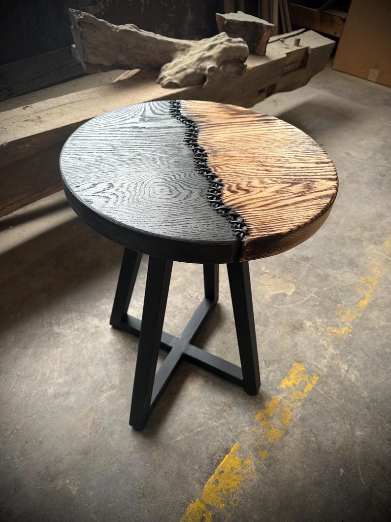 Op de afbeelding: Een ronde houten bijzettafel met een zwart en naturel houten blad. Het blad is in twee&euml;n gedeeld met een zwarte epoxy naad. De tafel heeft een zwart metalen driepoots onderstel.