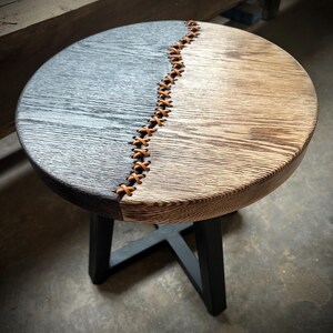 Op de afbeelding: Een ronde houten bijzettafel met een zwart en naturel houten blad. Het blad is in twee&euml;n gedeeld met een leren strook die in een kruispatroon is gestikt. De tafel heeft een zwart metalen onderstel.