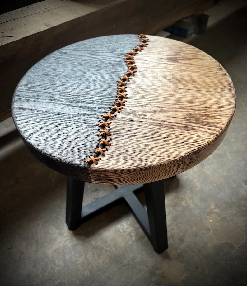Op de afbeelding: Een ronde houten bijzettafel met een zwart en naturel houten blad. Het blad is in twee&euml;n gedeeld met een leren strook die in een kruispatroon is gestikt. De tafel heeft een zwart metalen onderstel.