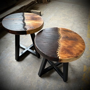 Op de afbeelding: Twee bijzettafels met zwarte metalen frames en houten bladen. De bladen zijn in twee&euml;n gedeeld, &eacute;&eacute;n kant is donkerbruin en de andere is lichtbruin hout. De twee helften worden verbonden door een decoratief stikdetail.