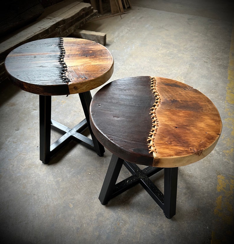 Op de afbeelding: Twee bijzettafels met zwarte metalen frames en houten bladen. De bladen zijn in twee&euml;n gedeeld, &eacute;&eacute;n kant is donkerbruin en de andere is lichtbruin hout. De twee helften worden verbonden door een decoratief stikdetail.