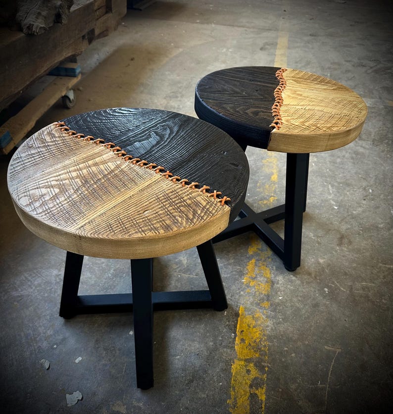 Op de afbeelding: Twee ronde houten bijzettafels met zwart gebeitst blad en natuurlijke houten onderkant. De bladen zijn in twee&euml;n gedeeld met een bruine leren naad. De tafels hebben zwarte metalen poten.