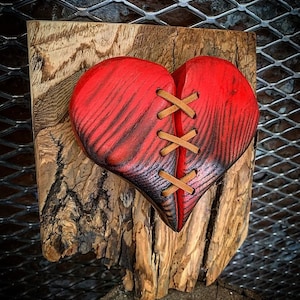 May include: A wooden heart sculpture with a red stain and leather stitching. The heart is broken in two and stitched together with brown leather thread.