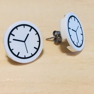 May include: A pair of white clock face earrings with black hands and a black outline. The earrings are on a light wood surface.