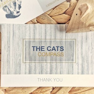 May include: A white thank you card with a wood grain pattern. The card has a gold frame with the text "THE CATS COMPASS" in blue lettering. The text "THANK YOU" is printed in gold lettering at the bottom of the card.