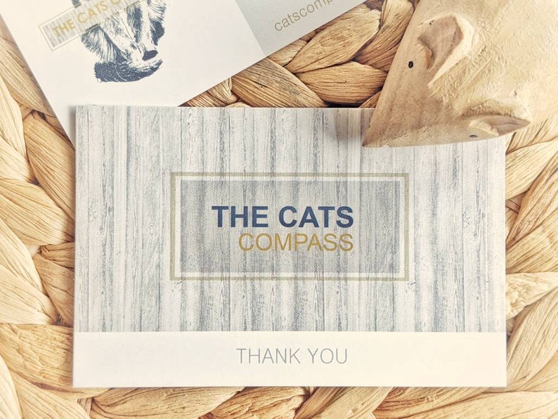 May include: A white thank you card with a wood grain pattern. The card has a gold frame with the text "THE CATS COMPASS" in blue lettering. The text "THANK YOU" is printed in gold lettering at the bottom of the card.