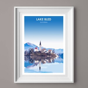 May include: A framed print of Lake Bled in Slovenia, featuring a picturesque view of the island church with its iconic steeple, surrounded by snow-capped mountains and a calm, reflective lake.