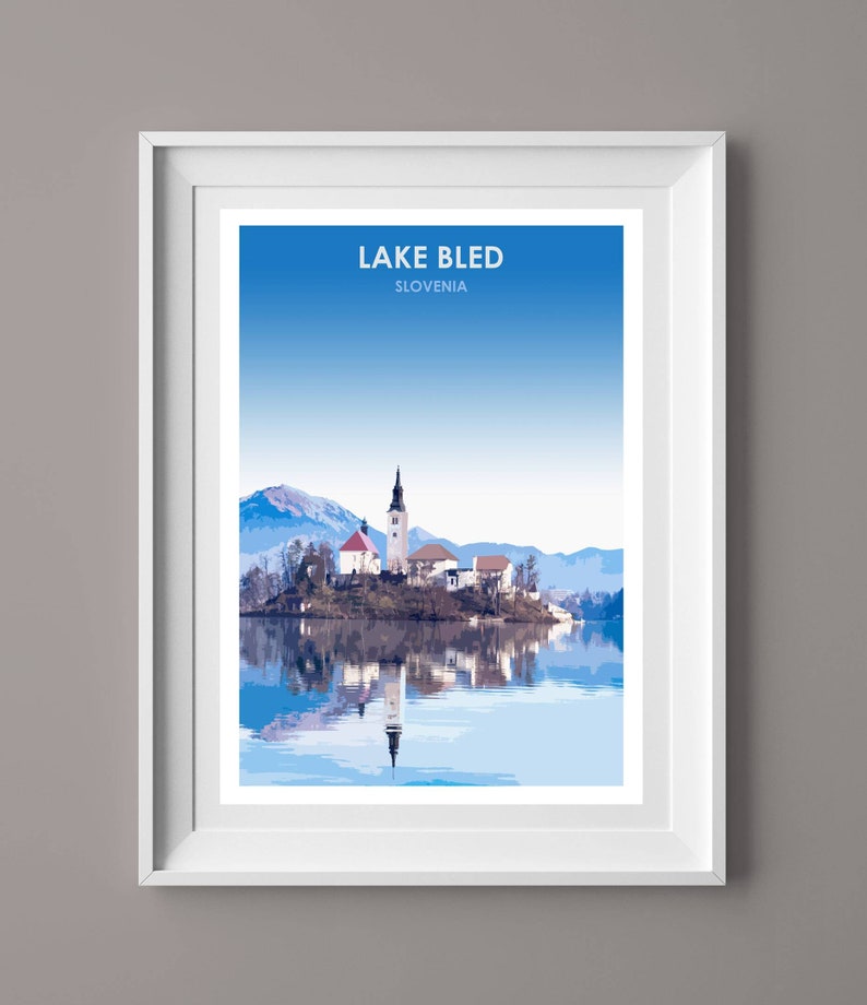 May include: A framed print of Lake Bled in Slovenia, featuring a picturesque view of the island church with its iconic steeple, surrounded by snow-capped mountains and a calm, reflective lake.
