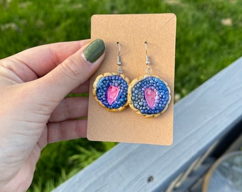 May include: A pair of earrings with a round, blue and pink design. The earrings are made of polymer clay and have a gold rim.