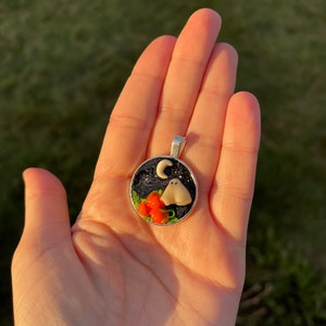 May include: A round silver pendant with a black background, a white ghost, a crescent moon, and orange pumpkins. The pendant is on a hand.