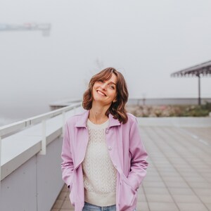 以下が含まれることがあります： 白いセーターの上に薄い紫色のジャケットを着て、薄いブルーのジーンズを履いた女性。彼女は笑顔で上を見ている。
