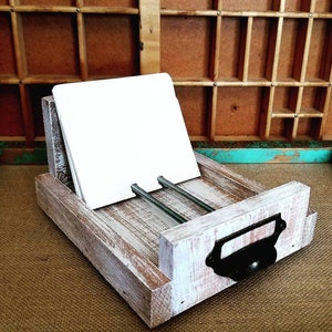 May include: A rustic wooden business card holder with a distressed white finish. The holder features a black metal handle and two metal rods to hold the cards. A stack of white cards is in the holder.