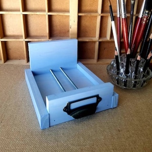 May include: A light blue wooden drawer with a black metal handle. The drawer is open and has two metal rods inside. The drawer is sitting on a brown surface.