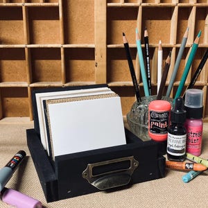 May include: A black wooden drawer with a brass handle holds white and tan cards. Pencils and art supplies, including paint and ink bottles, are arranged around the drawer. A wooden organizer with multiple compartments is in the background.