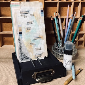 May include: A black wooden drawer with a brass handle holds a stack of altered book pages with text and painted accents. Colored pencils in a glass holder and a bottle of Distress Paint are nearby, along with two crayons.