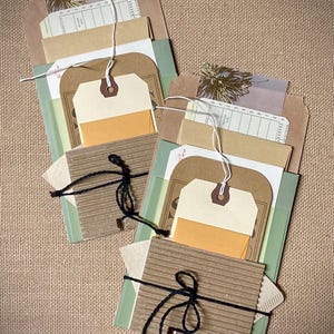 May include: Two handmade paper ephemera packs. Each pack includes a variety of vintage-style tags, receipts, and envelopes in shades of brown, cream, and green. The packs are tied with black twine and white string.