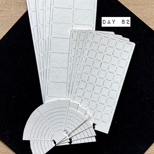 May include: A set of white paper planner stickers with different grid patterns. The stickers include a variety of grid sizes and shapes, including squares, rectangles, and circles. The stickers are labeled "DAY 82".