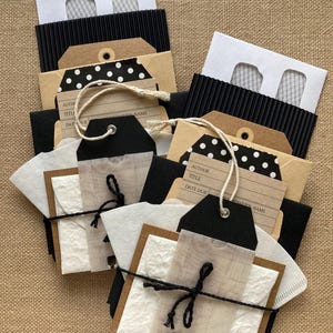 May include: A collection of handmade paper goods, including tags, envelopes, and decorative elements. The tags are black, brown, and white with text, and some have polka dots. The items are tied together with twine and black string.