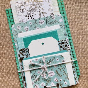 May include: A stack of stationery items, including cards and tags, with floral and botanical designs. The color palette includes white, teal, and gold. The items are tied together with white twine, creating a cohesive set.