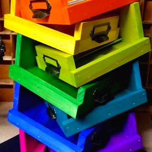 May include: A stack of colorful wooden storage boxes in a rainbow of hues: orange, yellow, green, blue, and purple. Each box has a metal handle and a drawer. The boxes are stacked at an angle, creating a visually appealing display.